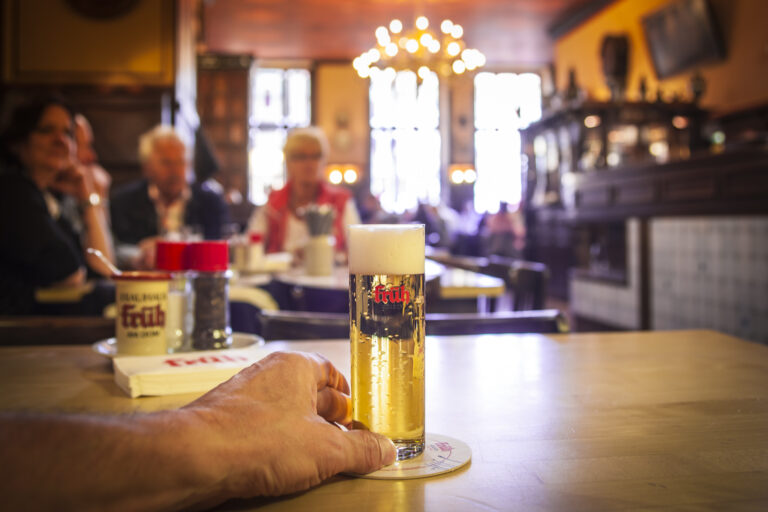 Eine Hand hält ein Glas Kölsch auf einem Holztisch in einer gemütlichen, warm beleuchteten Kneipe mit verschwommenen Gästen im Hintergrund und kunstvollem Dekor.