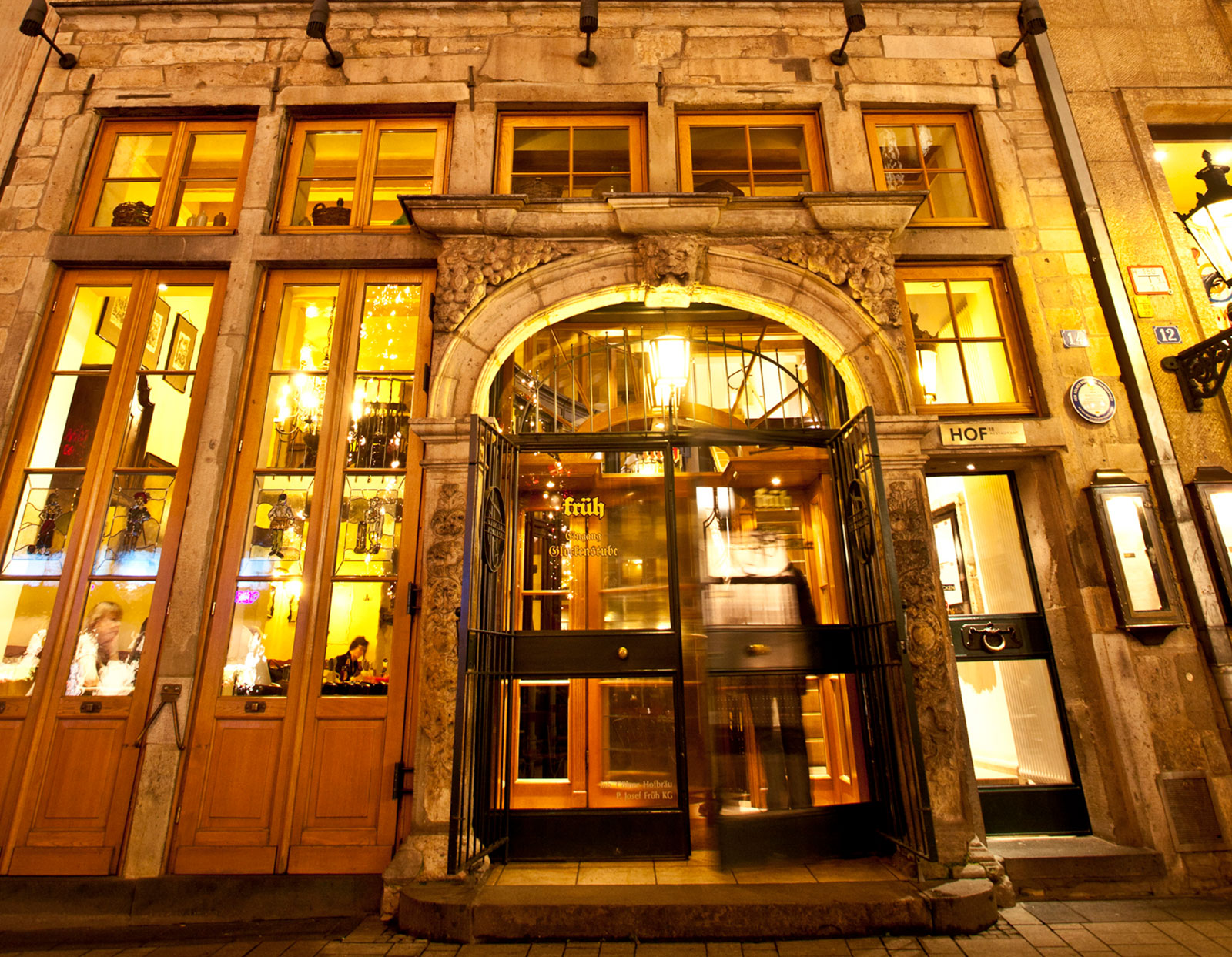 Goldgelbe Lichter beleuchten die Steinfassade und den kunstvollen Eingang eines historischen Gebäudes mit großen Fenstern, Menschen, die darin speisen, und einer verschwommenen Gestalt, die nachts durch eine Drehtür geht.