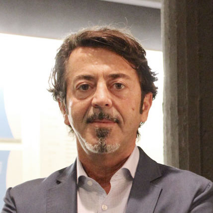 Ein Mann mit kurzen dunklen Haaren, Schnurrbart und Spitzbart, der ein blaues Jackett und ein helles Hemd trägt, steht mit neutralem Gesichtsausdruck im Haus.