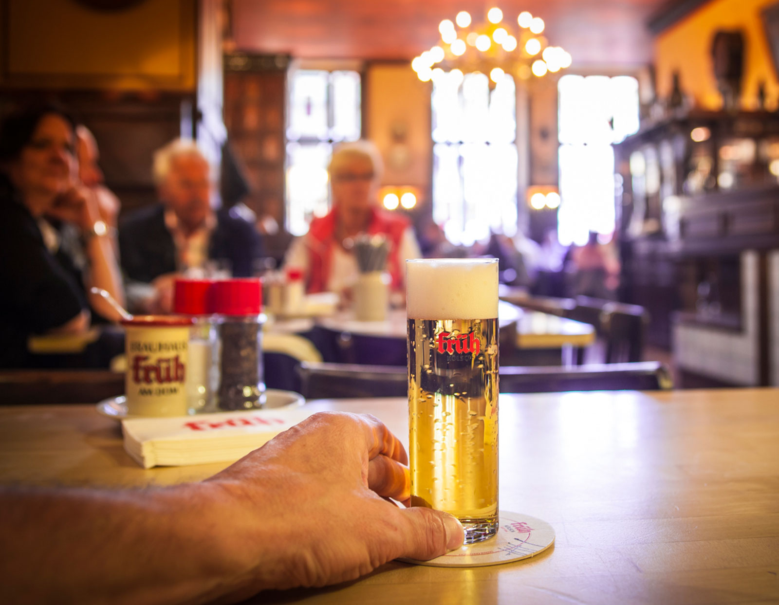 Eine Hand hält ein Glas Bier auf einem Tisch in einer gemütlichen, warm beleuchteten Kneipe, im unscharfen Hintergrund sitzen Leute und unterhalten sich.