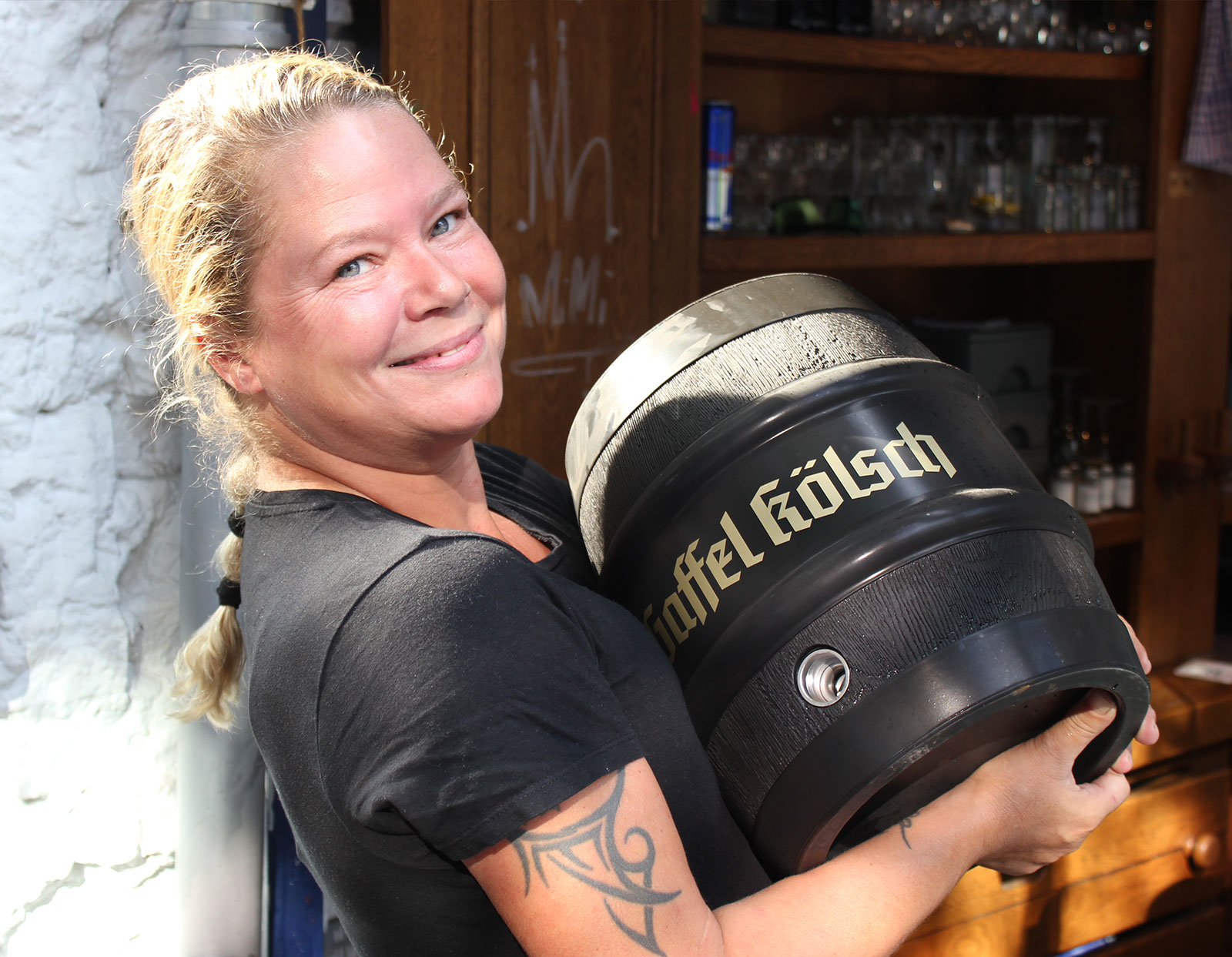 Eine lächelnde Frau mit einem Tattoo auf dem Arm hält während der Brauhaustouren in Köln ein schwarzes Fass mit der Aufschrift „Gaffel Kölsch“ in einem sonnigen Innenraum mit Regalen voller Gläser im Hintergrund.