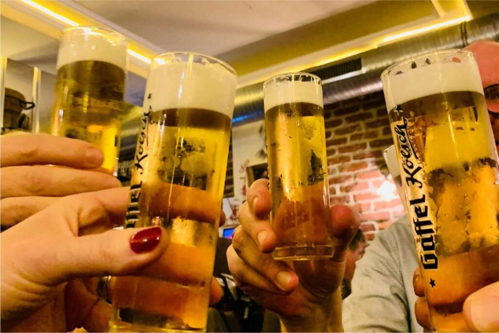 Mehrere Personen stoßen bei den Brauhaustouren Köln in einer Bar mit freiliegenden Backsteinwänden und warmer Beleuchtung mit hohen Biergläsern an und stoßen an.