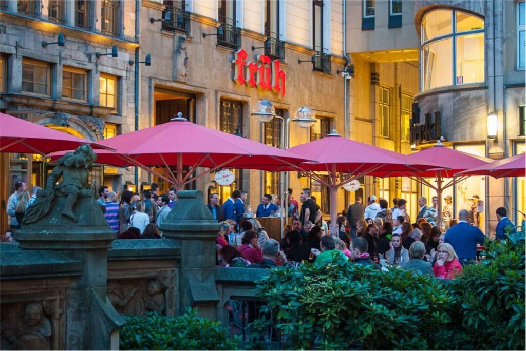 Ein lebhafter Biergarten im Freien mit vielen Menschen, die unter roten Sonnenschirmen sitzen und sich in der Abenddämmerung vor einem historischen Gebäude mit dem beleuchteten "Früh"-Schild treffen.