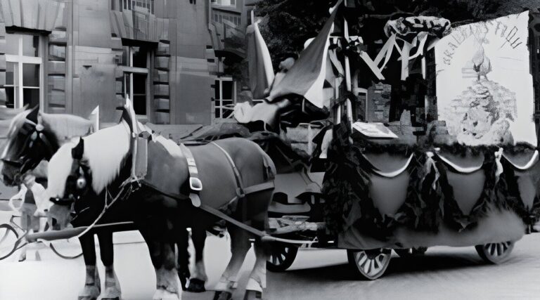 Zwei Pferde ziehen einen geschmückten Umzugswagen mit Grünzeug und einem großen, illustrierten Transparent. Der Wagen erscheint vor einem steinernen Gebäude. Die Szene hat ein festliches, historisches Thema.